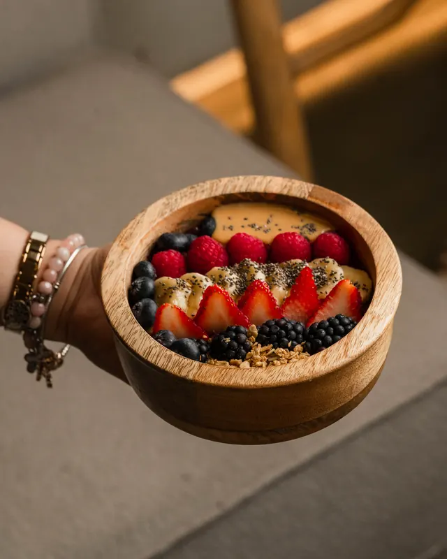 A hand holding a wooden açaí bowl topped with strawberries and blueberries